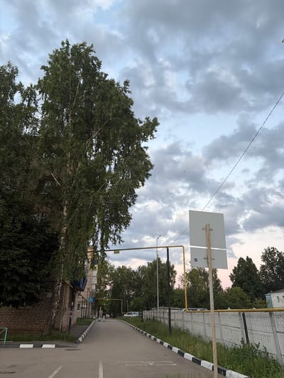 The image shows a street in the city of Ilchimbetovo, Russia, on a cloudy day. A tall birch tree stands to the left, with its branches extending over a brick building. A paved road runs through the center of the image, bordered by a concrete fence and a sidewalk. There are a few people walking on the sidewalk and a car parked in the distance. A white sign on a pole is visible on the right side of the road. The sky is filled with grey clouds, suggesting an overcast day.