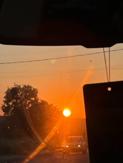 This media file depicts a sunset viewed through a window, likely from inside a vehicle. The sun, a bright, luminous orb, is positioned in the upper right quadrant of the image, casting a warm, orange glow across the scene. Its light is diffused by the haze or dust in the atmosphere, creating a soft, ethereal effect.
The foreground is dominated by a dark, reflective surface on the left, possibly the interior of the vehicle or a shadow cast by its structure. To the right of this, a light-colored frame or pillar of the window is visible, with thin wires running horizontally across it.
In the background, a tree with dark foliage is silhouetted against the vibrant sunset sky. The texture of the tree's branches and leaves is discernible, despite being backlit. The overall scene suggests a tranquil and possibly contemplative moment. The time of day is clearly sunset, indicated by the intense orange and yellow hues of the sky and the low angle of the sun. The presence of haze suggests it might be a warm, dry day, or potentially a day with some atmospheric pollution.
There are a few small, bright spots visible on the window surface, which could be reflections from within the vehicle or dirt/water droplets. The mood conveyed is one of peacefulness and the beauty of nature's daily spectacle. No text is visible in the image. The location context of Medovo, Bulgaria, adds a specific geographical element to this otherwise universal experience of observing a sunset.