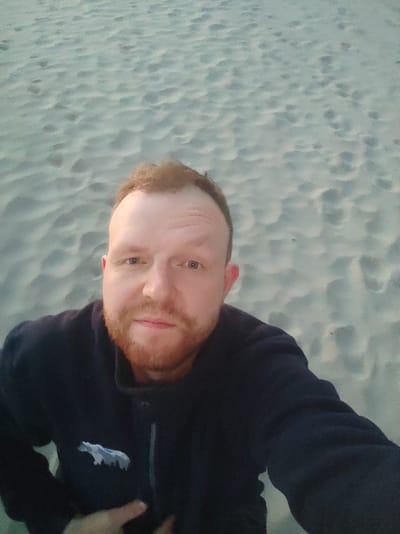 A man with red hair and a beard is taking a selfie on a sandy beach. He is wearing a dark blue jacket with a light blue embroidered polar bear on the left side of his chest. The background is a sandy beach with ripples and some footprints. The lighting suggests it may be dusk or dawn. The location of Motornoe, Russia is not identifiable from the image.