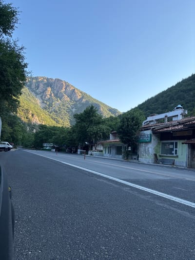 The image depicts a roadside scene in the city of Stara Kresna, Bulgaria. A paved road with white markings runs through the frame, flanked by a row of buildings and lush greenery. In the background, a majestic mountain range rises, its rugged slopes illuminated by the soft glow of sunlight. Trees densely cover the lower parts of the mountains, creating a stark contrast with the rocky peaks.
On the right side of the road, a collection of buildings appears to house businesses, indicated by signs. One building, painted in a faded green, displays a sign that reads "RAfting". Another sign above a storefront reads "КАФЕ РЕСТОРАНТ" (Cafe Restaurant). The buildings exhibit signs of age, with some appearing weathered and in need of repair. The architecture suggests a rustic, perhaps traditional style, with tiled roofs and stucco facades.
Several vehicles are visible. A white van and a dark-colored car are parked on the left side of the road. On the right side, near the buildings, a dark SUV is parked. Orange traffic cones are placed along the road. The overall atmosphere is one of a rural or semi-urban setting, characterized by a blend of natural beauty and small-scale commercial activity. The sky is clear and blue, suggesting fair weather.