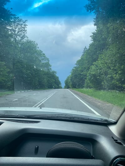 The image is taken from inside a vehicle, looking forward through the windshield. The scene depicts a paved two-lane road with white markings, curving slightly to the left as it recedes into the distance. Both sides of the road are lined with dense green trees. The sky above is overcast with dramatic, bluish-gray clouds, hinting at impending rain. The dashboard of the vehicle is visible in the foreground, with a dark textured surface. In the distance, a few road signs can be observed on the right side of the road. The overall environment suggests a rural setting in Belarus, possibly near Грозаўскі сельскі Савет.