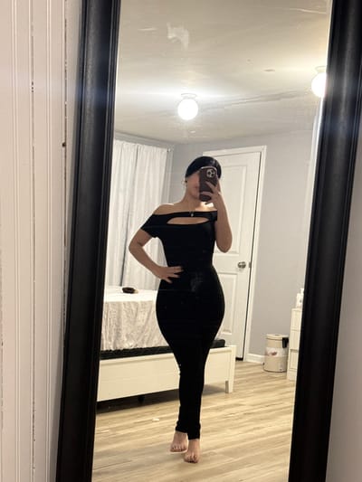 A person in a black, form-fitting jumpsuit takes a mirror selfie. The jumpsuit has short sleeves and a cut-out detail at the shoulder. They stand on a wooden floor with their left hand on their hip, holding a phone in their right. Behind them is a white bed frame and a door. Two round, wall-mounted lights illuminate the wall above. The selfie is taken in a large, ornate black-framed mirror. Below the mirror are white, horizontally-paneled walls.