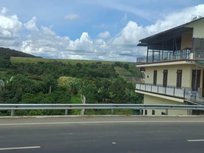 The image shows a roadside view in Đại Ninh, Vietnam, with a multi-story building to the right and a lush green hillside in the background. The building, a light yellow color with dark trim, features multiple windows, a balcony with white railings, and a metal roof extension. It appears to be a commercial establishment or residence. In front of the building and extending across the middle of the image is a gray metal guardrail separating the road from a grassy embankment. The road itself is asphalt with white lane markings visible. The background is dominated by a vast, green hillside covered in dense vegetation, with a partly cloudy blue sky above. Several power lines are strung across the sky in the middle ground. The overall impression is of a scenic rural or semi-rural location.