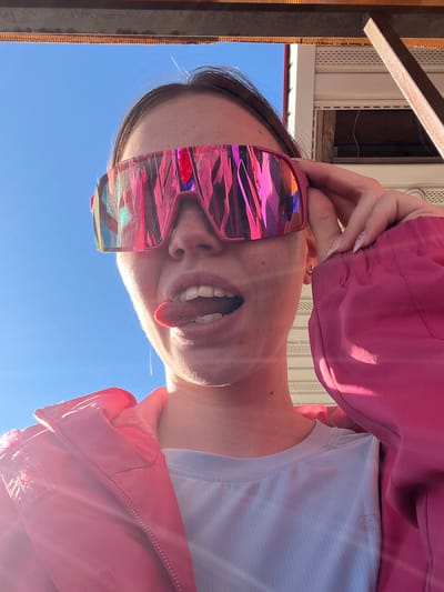 A young person is posing for a selfie outside in Gory, Russia. The subject is tilted at an angle, looking down at the camera with their tongue sticking out. They are wearing pink iridescent sunglasses and a pink jacket over a light blue shirt. The sky is blue and visible in the background. Part of a building structure is visible in the upper left corner. The setting appears to be a sunny day.