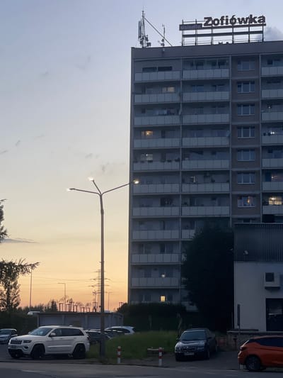 This media file depicts an urban scene in Jastrzębie-Zdrój, Poland, likely during the early evening as indicated by the soft, warm light of the sunset. The primary subject is a large, multi-story residential building with numerous balconies and windows, some of which are illuminated. A prominent sign is attached to the side of the building, displaying the text "Zofiówka" and "Osiedle" in large, dark letters.
In the foreground, a street lamp with three lights is visible, casting a subtle glow. The background reveals electrical pylons and transmission lines, silhouetted against the pastel hues of the sunset sky, suggesting the proximity of industrial or utility infrastructure. Several cars are parked along the street, including a white SUV in the foreground and other vehicles partially visible. The overall setting appears to be a residential area with a mix of buildings and open space, possibly a park or common area with trees. The weather seems calm, with a clear or lightly clouded sky. The image conveys a sense of everyday life in a Polish city during twilight.