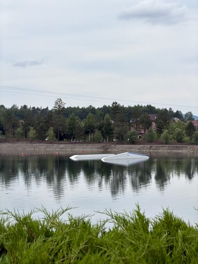 The image depicts a wake park located on a lake in the city of Озерное (майминский), Russia. In the foreground, lush green bushes line the bottom of the frame, partially obscuring the view. The lake itself is calm, with a smooth surface reflecting the overcast sky and the surrounding environment. Several orange buoys are visible in the water, marking a course or area.
Dominating the mid-ground of the lake are two white wakeboard ramps. One ramp is partially submerged, with its angled surface jutting out of the water, while the other is more fully visible, also angled. These structures suggest active use of the lake for water sports.
In the background, a treeline of various green trees forms a dense backdrop, with some houses and buildings nestled among the foliage, indicating a populated area bordering the park. Power lines are strung across the upper portion of the image, crossing the sky. The sky is filled with light clouds, suggesting a cool, possibly humid day. The overall scene is serene and suggests a recreational outdoor setting.