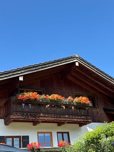 An exterior, low-angle, medium shot shows the upper section of a wooden chalet-style house against a clear blue sky. The roof is shingled with wood, and the upper facade is also made of dark, stained wood. A balcony with a dark wooden railing, intricately carved with geometric patterns, extends from the upper floor. Window boxes filled with vibrant orange and pink flowers adorn the balcony, and a smaller window with a green frame is visible above it. Below the balcony, a white-painted wall features two windows, one with dark shutters. Lush green foliage is visible in the lower right corner of the frame, partially obscuring another window with a pink flower box. The scene is likely set in Bischofswiesen, Germany, a region known for its traditional alpine architecture and floral displays.