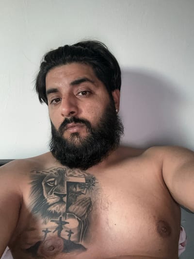 A man with dark hair and a beard is lying down, looking at the camera. He is shirtless, and has a tattoo on his chest. The tattoo depicts a lion's head, Jesus with praying hands, and three crosses on a hill. The background is a plain wall. The image was taken indoors, likely in a bedroom, in Mainburg, Germany.