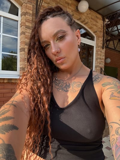 This is a vertically oriented, close-up photograph of a woman lying on her side, outdoors. She has long, wavy, reddish-brown hair with braids woven in. Her face is turned towards the camera, with her right eye in focus and her left eye partially obscured. She has noticeable makeup, including eyeshadow, and a nose ring. She is wearing a black, ribbed tank top and a silver necklace with a spherical pendant. Tattoos are visible on her arms and chest, some of which feature script. She is wearing a sparkly earring on her right ear.
The background shows a textured, light brown brick wall and a large window with reflections of the sky and trees. A white, globe-shaped light fixture is also visible on the wall. The lighting suggests it is daytime, with strong sunlight casting shadows, indicating fair weather. The overall impression is one of a casual, relaxed moment captured outdoors. There is no visible text in the image.