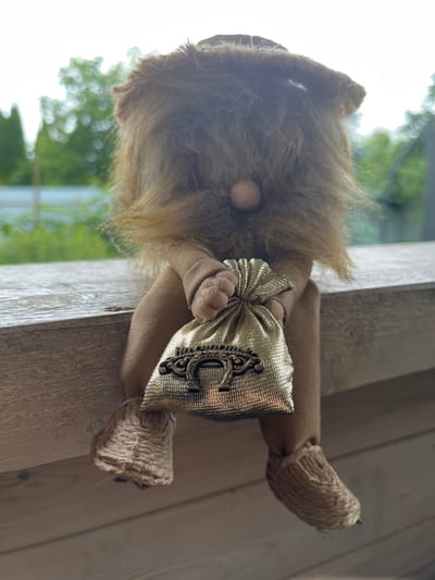 A stuffed gnome figure with a brown beard and a tan body is sitting on a wooden railing. The gnome is holding a small, shimmering gold pouch with a decorative metal emblem on it. The background shows blurred green trees and a bright sky, suggesting an outdoor setting. The gnome's feet are made of rough, woven material.