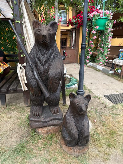 The scene depicts two carved wooden bear sculptures positioned in an outdoor setting. The larger bear stands upright, holding a long, narrow object, potentially a rifle, across its chest. The sculpture is resting on a rectangular base, which appears to be part of a wooden bench. A white cloth is visible next to the bear on the bench. A smaller bear sculpture sits nearby on a cylindrical wooden base, positioned directly on the grass. A black metal post stands behind the two bears. In the background, a building with yellow walls and brown doors can be seen, partially obscured by lush greenery and pink and red flowers. The ground is covered in dry grass and patches of dirt. The scene appears to be located in Goliam Dol, Bulgaria, based on the contextual information.