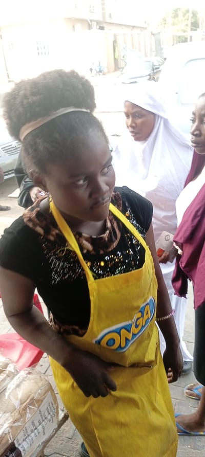 A woman wearing a bright yellow apron with "ONGA" printed on it stands with two other women. She has dark skin and wears her hair up with a headband. The woman to her right is wearing a white hijab. A third woman stands to the side, partially visible. In the background, there's a glimpse of a street scene with cars and buildings. Some items, possibly food products, are visible near the front of the frame.