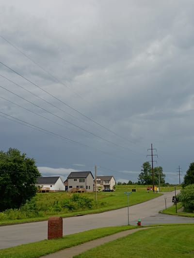 The image depicts a street intersection in a suburban area of Paris, United States of America. The sky is overcast with heavy, dark clouds. Several power lines stretch diagonally across the frame from the left to the right. In the middle ground, a paved road curves through a grassy landscape dotted with young trees and bushes. Three houses are visible on a slight incline to the left and center of the road, each with dark roofs. One house is white with a wooden deck, another is gray with a similar deck and black window frames, and a third is white with a red-brick chimney visible. A brick mailbox stands near the foreground on the left side of the road. A green and red fire hydrant is located on the right side of the road at the intersection. A street sign reading "MILLSTONE" is visible. Two tall, dark utility poles with crossbars are positioned in the background. The overall scene suggests a residential neighborhood on a cloudy day.