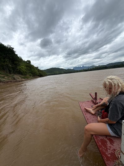 The image captures a wide shot of a brown river with a cloudy sky overhead. A person with blonde, curly hair is sitting on the edge of a red boat, with their legs dangling over the side and one foot in the water. The boat appears to be on a river, with lush green hills lining the opposite bank and in the background. The scene is set in Thinhong, Laos, as indicated by the provided context. The overall atmosphere is serene and natural.