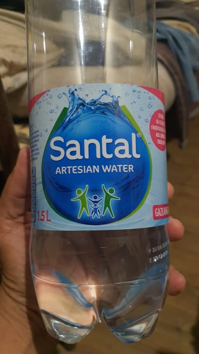 The image shows a close-up of a plastic bottle of "Santal Artesian Water". The bottle is partially filled with water. A person's hand is holding the bottle. The label on the bottle is primarily blue and white, featuring a water splash graphic and stylized figures of people. Text on the label includes "Santal", "ARTESIAN WATER", and "1.5L". There is also some text in a foreign language on the upper right side of the label, and a red "GAZLAN" logo on the lower right. Below the main label, there is printed text on the bottle itself, including "P 30/06/2026" and "E 27/12/2020". The background is out of focus, showing hints of fabric and wood. There is no indication of the city of Karankul, Uzbekistan within the image itself.