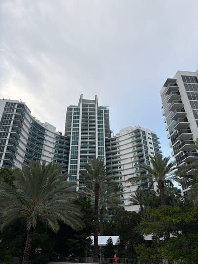 The image shows a low-angle view of several modern, white high-rise buildings with numerous balconies and windows, interspersed with lush green palm trees and other foliage. The architecture features clean lines and a repetitive, grid-like structure for most of the buildings, though one building exhibits a curved, organic design. The sky above is overcast and appears pale blue or grey, suggesting it might be daytime with cloudy weather.
The scene depicts an urban environment, likely a coastal city, given the presence of palm trees and the style of architecture often associated with warm climates. There are no people visible in the image, and the focus is on the buildings and natural elements. The overall impression is one of modern development integrated with tropical vegetation.
A very faint text label on one of the curved buildings appears to read "CANTOR COLLINS" followed by more unreadable text, potentially indicating the developer or architect. The image is oriented vertically, with the buildings extending upwards into the sky. The lighting suggests it is likely morning or late afternoon, with diffused light due to the cloud cover. The setting appears tranquil, with no immediate signs of activity other than the static presence of the structures and trees.