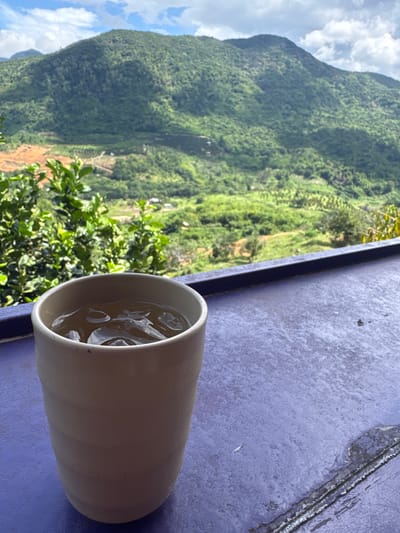 A close-up shot features a light-colored ceramic cup filled with ice and a light-colored beverage. The cup sits on a textured purple surface. In the background, lush green mountains with some terraced fields and scattered structures dominate the view under a cloudy sky. The overall scene suggests a tranquil, possibly rural, setting.