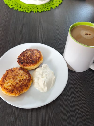 A close-up, high-angle shot captures a breakfast scene on a dark wood-grained table. In the foreground, a white plate holds two golden-brown syrniki (cottage cheese pancakes) and a dollop of white sour cream. To the right, a white mug with a green rim contains coffee with a light brown crema, exhibiting a small swirl of darker foam on top. A bright green doily is partially visible in the upper left corner. The setting is domestic, suggesting a home environment in Brodi, Latvia. There are no people or actions depicted.
