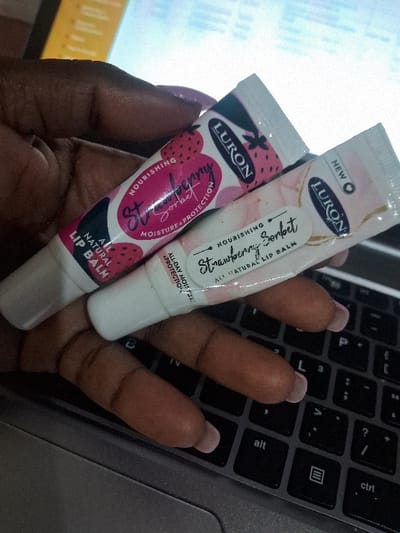 The image shows two tubes of Lurón brand lip balm. The smaller tube, labeled "Strawberry," is mostly pink with illustrations of strawberries. The larger tube, labeled "Strawberry Sorbet," has a pink and white marbled background. Both tubes claim to be "all-natural lip balm" and promise "nourishing moisture and protection." They are held in a hand with manicured nails, positioned over a gray laptop keyboard.