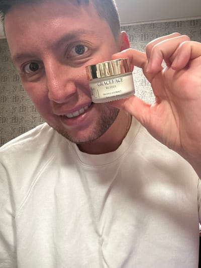 A man holds a small jar of "Graceface Russia Truffle Extract Face cream, 30g" close to his face. The background consists of a wall with a repeating pattern of the words "STOCK", "MARKET", and "FRAU." The man is wearing a white sweater. The location, based on the cream's label, is likely Vorotynsk, Russia.