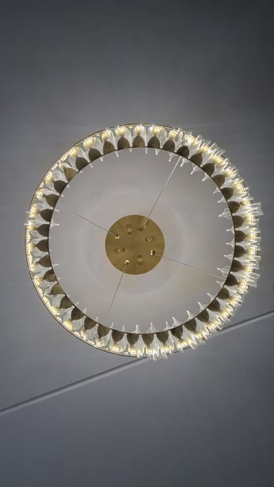 A close-up, worm's-eye view shows a circular chandelier hanging from a light gray ceiling. The chandelier has a gold-toned metal frame and is adorned with numerous clear, pointed glass elements that are lit from within, giving off a warm, yellow light. The chandelier is suspended by thin wires connected to a central gold-toned circular canopy attached to the ceiling. The ceiling itself has a subtle texture and a faint diagonal line across its surface. No people or other objects are visible in the scene. The location is specified as Верзилово, Russia.
