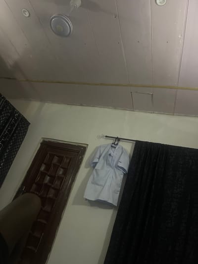 In Pantinapu, Nigeria, this image captures an indoor scene with a ceiling fan and recessed lighting overhead. A white, short-sleeved shirt with blue trim hangs on a rod, adjacent to a dark, patterned curtain. To the left, a dark brown door with a textured panel is visible. A dark, indistinct object, possibly a limb, intrudes into the lower left corner of the frame. The room appears dimly lit with a predominantly white wall and a light-colored, paneled ceiling.