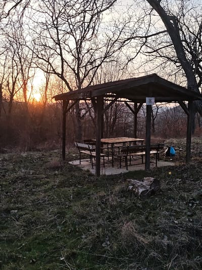 The image displays an outdoor picnic shelter situated in the city of Opaka, Bulgaria, observed at either sunrise or sunset. The central subject is a dark-stained wooden gazebo featuring a gabled roof supported by six visible posts, enclosing a rectangular wooden picnic table with integrated benches on all four sides. The structure rests upon a light-colored concrete slab. A small, rectangular white sign, bearing an indistinct dark symbol, is affixed to one of the gazebo's interior support posts. A blue and black bag or backpack is present on the ground immediately to the right of the gazebo. The foreground is composed of sparse, dry grass, fallen leaves, and areas of bare earth. A horizontal tree log or stump is positioned in the lower right. The background consists of numerous deciduous trees, devoid of foliage, silhouetted against a pale sky. The sun, partially obscured by trees, is visible on the left horizon, emitting a pronounced orange glow. Distant undulating terrain is faintly discernible through the bare tree branches. No individuals or active interactions are depicted in the scene.