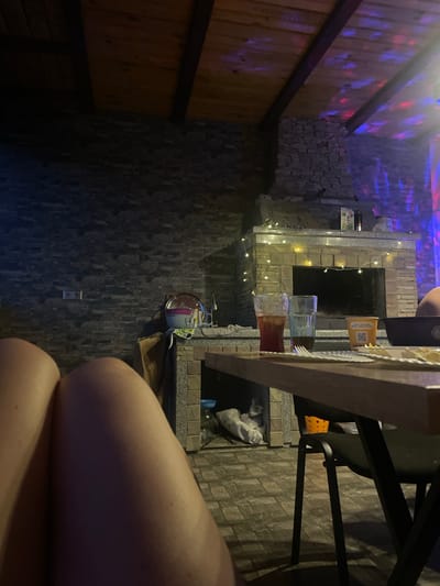 The image captures a dimly lit indoor or semi-enclosed outdoor area in Petrivka, Ukraine, from a low perspective, likely of a person seated at a table.
In the foreground, the bare upper thighs of a person occupy the lower left and central portions of the frame. A wooden table extends into the scene from the lower right. On the table, from left to right, are two tall glasses, one containing a reddish-brown liquid with ice, and another containing a darker liquid. A yellow disposable cup with Ukrainian text reading "ЇЖА З ДОСТАВКОЮ ЧЕРКАСИ" (Food with delivery Cherkasy) is visible, along with a black bowl and a white scalloped-edge plate or napkin with a fork resting on it.
The midground features a prominent stone-tiled wall on the left, equipped with a double electrical outlet. A substantial built-in structure, appearing as a fireplace or grill unit, dominates the central area. It is constructed from light-colored stone and features a hearth. A string of warm white decorative lights is draped across its mantelpiece. Resting on the mantelpiece are a small potted item, a dark bottle, and a glass jar. To the immediate left of this structure, a small countertop with a sink, a large clear bowl, a smaller purple bowl, and a white cloth adorned with an avocado pattern are visible. Beneath the table, a black metal chair stands, with an orange plastic basket positioned beneath it. A bundle of white material rests on the stone floor to the left of the chair.
The background reveals a wooden plank ceiling supported by dark wooden beams. The ceiling and upper right wall are illuminated by dynamic red, blue, and purple colored lights, casting reflective patterns across the surfaces. The walls throughout the space are faced with dark grey or brown rectangular stone tiles.