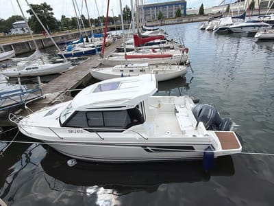 The image depicts a white motorboat with the registration "SW-J 610" docked in a harbor. The boat has a cabin with tinted windows and a Yamaha outboard motor at the stern. It is moored next to a wooden pier, with other sailboats visible in the background. The harbor is located in Wismar, Germany, a Hanseatic city known for its historic port. The sky is overcast, and the water is calm. There are no people visible in the image. The boat appears to be in good condition and is ready for use.