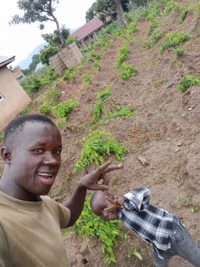The image captures a selfie taken outdoors in what appears to be a rural area, possibly in Baltap, Nigeria. In the foreground, a man is smiling and looking towards the camera, with his left hand raised and extended towards another individual. To his right and slightly behind him, a young boy is also smiling and making a peace sign with his right hand. The background shows a sloped, cultivated field with rows of young green plants. Several buildings, some appearing to be houses with corrugated metal roofs, are visible further up the slope. A large tree stands prominently in the mid-ground. The ground is a mixture of soil and dry vegetation. The sky is overcast.