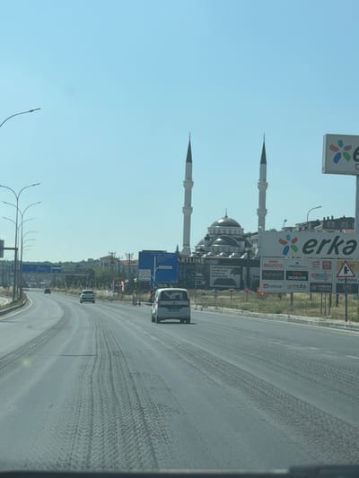 This is a medium-distance, eye-level, outdoor, daytime shot of a multi-lane road in Kütahya Merkez, Turkey. The road appears to be paved with asphalt and shows tire tracks from vehicles. A white car is visible in the right lane, and another white car is in the left lane. A mosque with two minarets is located in the background, to the left of a large billboard with advertisements. The sky is clear and blue. The scene appears to be a commercial or industrial area, given the presence of billboards and the architecture.