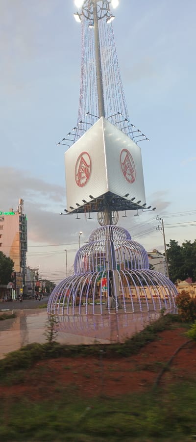 The image captures a brightly lit, modern city decoration in Buon Ho, Vietnam, likely at dusk or dawn. The central object is a tall, metallic structure resembling a Christmas tree, adorned with cascading strings of colorful lights. At its mid-section, a large, cube-shaped billboard with a prominent red and white circular logo on each visible face sits atop a decorative, layered base made of curved metallic frames, also outlined with blue lights. This base resembles an ornate birdcage or an abstract tiered fountain.

The background reveals a typical urban street scene in Vietnam. To the left, a multi-story building with a sign displaying "Hotel" in green neon lights stands prominently, alongside other commercial establishments with various signs. The street is paved and appears wet, reflecting the lights from the surrounding environment. A few motorcycles are visible on the street, suggesting moderate traffic. Power lines crisscross the sky, a common sight in urban areas. The sky is partly cloudy with hues of gray and pale blue, indicating either early morning or late evening. The foreground features a patch of reddish-brown soil and green vegetation, suggesting the decoration is situated in a traffic island or public square. The overall impression is one of a lively, decorated urban space transitioning between day and night.