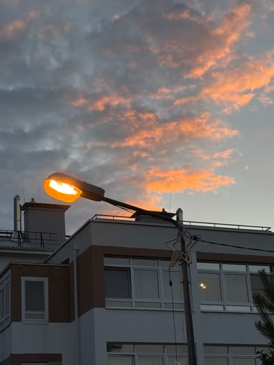 The image captures a streetlamp emitting a bright orange glow against a cloudy sky tinged with sunset colors. The streetlamp is mounted on a pole that also features a star-shaped decoration. In the background, a building with several windows is visible, partially obscured by the streetlamp and a tree branch on the right. The building appears to be a modern structure with a white and brown facade. The sky displays a mix of dark and light clouds, with patches of orange and pink suggesting the setting sun. The scene is set in the city of Michurinsky, Russia. There are no people visible in the image.
