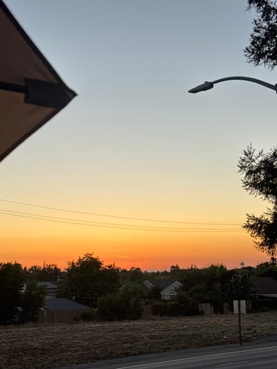 The image captures a residential neighborhood in North Highlands, California, at dusk. The sky exhibits a gradient of colors, transitioning from a pale blue at the top to a vibrant orange near the horizon, indicating the setting sun.
In the foreground, a patch of dry, mowed grass stretches across a slight incline. A paved road with a white line marking is visible at the bottom edge of the frame. A rustic, weathered signpost stands in the middle ground, to the right of center.
The middle ground is dominated by mature trees, interspersed with residential homes. The roofs of several houses are visible, and a wooden fence runs along the property line of at least one dwelling. Power lines are strung across the scene, supported by unseen poles.
The background features more trees and the distant silhouette of a water tower. A streetlamp with a curved arm is positioned in the upper right quadrant of the image, silhouetted against the twilight sky. A portion of a large, dark-colored umbrella canopy is visible in the upper left corner, suggesting the vantage point is from an outdoor seating area. No people or animals are present in the frame.