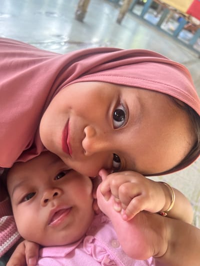 A diagonal close-up image depicts two individuals, an older child and an infant, in an indoor setting in Gembong, Indonesia. The older child, appearing to be a young girl, occupies the upper right frame, wearing a pink hijab and a matching top. Her dark eyes are open, and her lips, adorned with pink lipstick, are slightly parted in a subtle smile. Her right hand gently grasps the left foot of the infant, who is positioned below and to the left, wearing a light pink ruffled collared shirt. The infant looks towards the viewer with parted lips, and a gold-colored bangle is visible on its right wrist. The background is blurred, showing light-toned flooring and indistinct structural elements, indicating a well-lit interior.