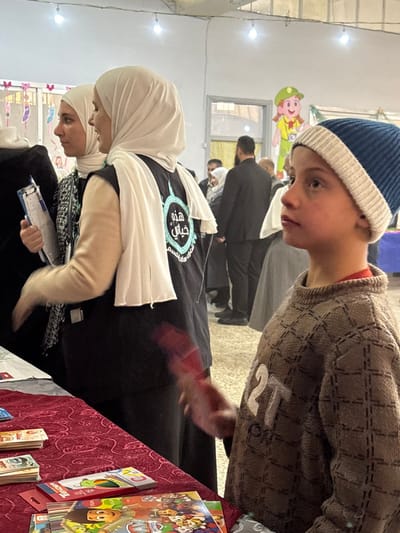 This image depicts an indoor scene in Kafr Batna, Syria, at what appears to be a community event or exhibition. In the foreground, a young boy is visible from the chest up, wearing a blue and white knit beanie and a brown patterned sweater with "2T" embroidered in white. He faces right in profile, looking upwards and right with a neutral expression.
To the boy's left, a table is covered with a red, intricately patterned tablecloth. On the table are several stacks of Syrian Pound banknotes, children's activity books (including one featuring "Paw Patrol"), and a box of "Tender" colored pencils.
Behind the table, two women are partially visible. The woman on the left wears a light-colored hijab and a black and white patterned top, holding a blue clipboard with papers; her right arm is slightly blurred, indicating motion. The woman to her right wears a white hijab and a black vest adorned with a light blue circular logo featuring Arabic text, specifically "هذه حياتي" (This is my life). Her left arm has a light beige sleeve, and her hand is blurred while holding a red object.
In the midground and background, multiple adults are present, some in dark suits, others in traditional attire including hijabs. A large cartoon cutout of a girl in a green hat and yellow top, holding a camera, is affixed to a wall in the background near a doorway. The indoor space has light-colored walls, a light-toned floor, and is illuminated by several bare light bulbs hanging from the ceiling. Colorful paper cutouts decorate a wall in the upper left background.