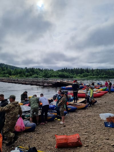 The image depicts a group of people preparing for a rafting trip on a river. The scene is set on a rocky shoreline with a forest-covered hill in the background. Several inflatable rafts are positioned on the shore, some of which are red, blue, and yellow. A wooden pontoon bridge extends across the river, with people visible on it. The sky is overcast with the sun partially visible through the clouds. The people are dressed in casual clothing, including camouflage attire and t-shirts. Some individuals are carrying bags, and others appear to be organizing gear. The overall atmosphere suggests the beginning of an outdoor adventure. The location is identified as Vishegorsk, Russia.