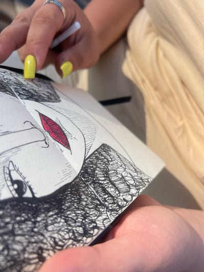 The image shows a close-up of a person's hands holding a sketchbook. The sketchbook contains a drawing of a face with detailed black ink lines and a red accent on the lips. The person's fingernails are painted bright yellow, and they are wearing a silver ring. One hand holds a white stylus or pen, poised as if to continue drawing. The background is blurred, showing a light-colored fabric, possibly clothing, and a hint of a shadow, suggesting an outdoor setting. The lighting appears to be natural, possibly indicating daytime. The focus is on the act of drawing and the partially completed artwork.
