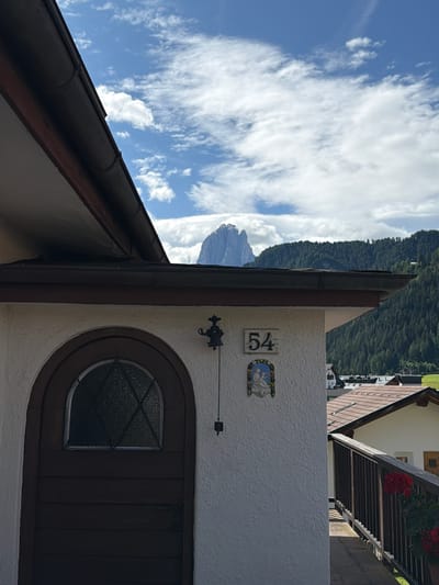 The image captures a view of a building in Urtijëi - St. Ulrich in Gröden - Ortisei, Italy, against a backdrop of mountains and a partly cloudy sky. The foreground features the side of a light-colored stucco building with a dark brown tiled roof and a dark brown arched wooden door. Above the door, a small, decorative bell hangs from a wire. To the right of the door, a ceramic plaque with the number "54" is mounted on the wall, and below it, another decorative ceramic plaque depicts a religious scene. A dark wooden railing runs along a pathway to the right of the building, with red flowers in a pot visible. In the background, lush green, tree-covered mountains rise, and a prominent, jagged mountain peak is visible in the distance under a blue sky with scattered white clouds. The architecture suggests a traditional alpine style.