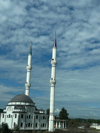 The image depicts a mosque located in the city of Menteşe, Turkey. The central structure is a multi-domed mosque, primarily constructed from light-colored stone or stucco, featuring a large, dark grey central dome and several smaller domes. The main building exhibits multiple arched windows and decorative architectural elements. Two tall, slender white minarets flank the mosque. Each minaret possesses at least two distinct balconies (şerefe) with blue geometric patterns, topped by a pointed spire with blue and white accents and a finial. A faint, net-like or decorative string of elements connects the upper sections of the two minarets. The scene is set under a partly cloudy blue sky with scattered white cumulus clouds. The foreground and mid-ground contain sparse green foliage and utility lines. To the right of the mosque, a smaller white structure with columns is visible, alongside additional trees and distant landforms or structures in the background.