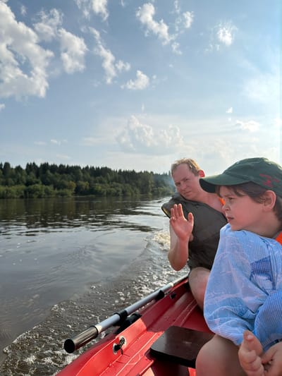 In the city of Kalino, Russia, two individuals are traveling in a red boat on a body of water, likely a river, based on the presence of a forested shoreline. The man in the background, wearing a dark vest and life jacket, is looking towards the camera with his hand raised in a waving gesture. In the foreground, a child wearing a green cap and a blue shirt, also with an orange life jacket visible, is looking away from the camera. The boat is moving, as indicated by the wake trailing behind it. The sky above is a pale blue with scattered white clouds. The trees on the opposite bank are dense and green, suggesting a natural, possibly rural, setting. The water is dark and slightly rippled. The overall scene depicts a leisurely boat ride on a sunny day.