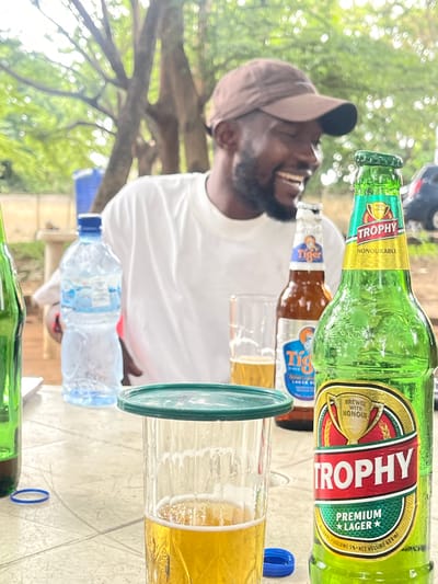The image shows a close-up of a man wearing a brown baseball cap and a white t-shirt. He is smiling and appears to be laughing. He is holding a green bottle of Trophy Premium Lager beer. In the background, there are other bottles and glasses, some containing beer. A bottle of water is also visible. The setting appears to be outdoors, with trees and foliage in the background, suggesting a casual gathering or celebration. The lighting suggests it is daytime. The Trophy bottle has the brand name "TROPHY" prominently displayed, along with "PREMIUM LAGER" and "BREWED WITH HONOUR". Another visible bottle appears to be a "Tiger Lager". The overall mood seems jovial and relaxed. The context provided indicates the location is Wupa, Nigeria.