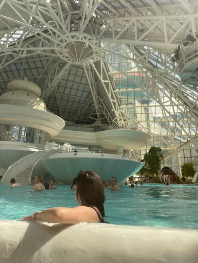 The image depicts people in a pool within the Caldea spa complex, located in Les Escaldes, Andorra. The distinctive architectural feature is the glass and metal structure forming the roof and walls of the building, which is visible above the pool. People are seen swimming in the pool, with one individual closer to the camera partially submerged in the water. The pool appears to be integrated with the building's structure, with white, rounded elements visible around the water's edge. The environment suggests an indoor, controlled climate spa setting.