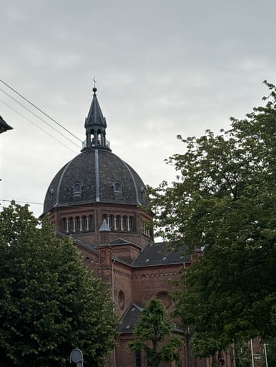 The image shows a close-up, low-angle shot of a domed brick building, likely a church, with a spire topping its dome. The building is partially obscured by the foliage of several large, leafy green trees in the foreground and right side of the frame. The sky is overcast and grey. Power lines are visible crisscrossing the upper left portion of the image. The architectural style of the building suggests it is an older structure, possibly from the late 19th or early 20th century. Given the context of Frederiksberg, Denmark, this building strongly resembles the historic Frederiksberg Church, also known as Kirken ved Sankt Thomas, located on Godthåbsvej. The building's brickwork, the arched windows visible beneath the dome, and the characteristic dome shape are consistent with this landmark. There are no people visible in the image. The overall scene conveys a sense of quiet solemnity due to the overcast sky and the presence of mature trees framing the edifice.