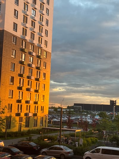 This media file shows an outdoor scene at dusk. The sky is filled with dramatic clouds, transitioning from a muted grey to warmer hues of orange and yellow as the sun sets.
Dominating the foreground is the facade of a modern apartment building, its windows reflecting the fading light. The building's exterior appears to be clad in a mix of light and dark textured tiles. Many of the windows are small and rectangular, some with visible interior structures or reflections. The building stretches across the frame, suggesting a residential complex.
Behind the apartment building, a large parking lot is visible, filled with numerous cars. The cars are parked in rows, and their variety in shape and color adds to the sense of a busy urban or suburban environment. Some trees and greenery are interspersed within the parking area and along the edge of the scene, with some deciduous trees showing red foliage, possibly indicating autumn.
To the right of the parking lot, a white van is parked, its side facing the viewer. Further down, several other cars are parked, including a dark-colored sedan and what appear to be SUVs and hatchbacks. The overall impression is one of a residential area with ample parking.
The lighting suggests it is either sunrise or sunset. Given the warm tones in the sky and on the building, sunset is more likely. The presence of cars and buildings points towards a developed area. No people are clearly identifiable as engaging in specific activities, though there might be figures near the parked cars or within the buildings that are not distinct enough to describe. The scene conveys a sense of the end of the day in a populated, modern setting. No visible text is present in the image.