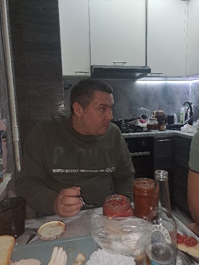 A man wearing a green sweater sits at a table laden with food. There are jars of tomato sauce, a clear glass bottle, and a plastic bag. The table holds bread, what appears to be pieces of fat, and other unidentifiable items. The background shows white cabinets and a dark backsplash. The image has been rotated counterclockwise.