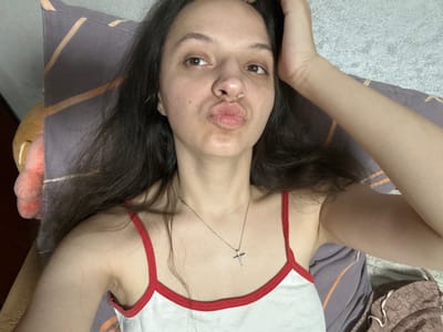 A young woman with long, dark hair is lying on her back, looking up at the camera. She is wearing a white tank top with red trim and a silver cross necklace. Her right hand is raised, touching her hair. Her lips are pursed in a kiss. She is lying on a bed with a pillow that has a brown and gold striped pattern. A stuffed animal is visible to her left. The scene is indoors, possibly in a bedroom. The location is not specified to be Postoyata, Ukraine.