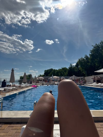 The image depicts a person's legs in the foreground, positioned on a lounger, looking towards a swimming pool area. The pool is filled with blue water and has a tiled edge. Beyond the pool, there are numerous lounge chairs and umbrellas. In the background, trees and foliage are visible, as well as a glimpse of the sky with white clouds and bright sunlight. The scene appears to be a resort or recreational area, possibly at a location near Vodnik, Russia, given the context. Several individuals are sparsely scattered around the pool area, some sitting on loungers and others in the pool.