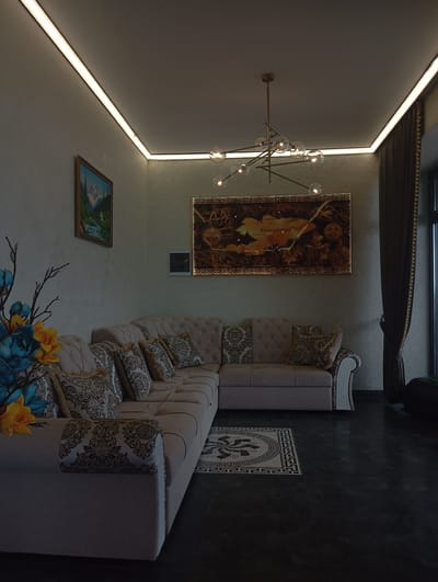 The image shows a well-lit modern living room with a sectional sofa, artwork on the wall, and decorative elements. The room features a sleek, contemporary design with a focus on indirect lighting along the ceiling.
The primary subject is a large, light-colored sectional sofa with tufted cushions and patterned throw pillows. The sofa is arranged in an L-shape and positioned on a dark, reflective floor. A decorative rug with a geometric pattern is placed in front of the sofa.
On the wall above the sofa, there are two framed pieces of artwork. The larger piece, centrally located, appears to be a map or a stylized depiction of Kyrgyzstan, with Cyrillic text visible. To the left of this, a smaller, framed painting depicts a mountainous landscape with a waterfall.
To the left of the sofa, a floral arrangement with blue and yellow artificial flowers is visible, adding a touch of color to the scene. The walls have a textured finish, and the ceiling is flat with integrated LED strip lighting, creating a soft, ambient glow.
A modern chandelier with multiple glass globes hangs from the ceiling over the seating area. To the right, dark curtains are drawn across a window, and a sliver of the window frame and exterior is visible.
The overall impression is of a sophisticated and comfortable living space. The location is identified as Бактуу-Долоноту, Kyrgyzstan, suggesting this is likely a residential interior within that area. There are no people visible in the image.