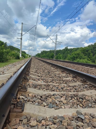 The image captures a low-angle view of a railway track extending into the distance, flanked by lush green trees on either side. The sky above is a vibrant blue, dotted with fluffy white clouds, suggesting a pleasant day. Overhead, complex electrical wires for the train are strung between tall, concrete poles, indicating this is an electrified railway line. The track itself is laid on a bed of ballast, composed of numerous small stones and resting on concrete sleepers. The rails are secured to the sleepers with metal clamps and bolts, showcasing the robust construction of the railway infrastructure.
The scene is devoid of people or any immediate activity. The focus is solely on the railway line and its surrounding environment. The sunlight casts no strong shadows, and the overall lighting is even, suggesting it could be late morning or early afternoon. The dense foliage of the trees implies a rural or semi-rural setting, far from a bustling city. The clear sky and abundant greenery evoke a sense of tranquility and the vastness of nature coexisting with human engineering.
There is no visible text in the image. The overall impression is one of a functional, yet serene, transportation artery set against a backdrop of natural beauty. The composition draws the viewer's eye along the converging lines of the tracks towards the horizon, emphasizing the journey and the endless possibilities it represents.